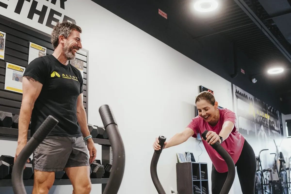 Two members sharing a laugh on the air bikes at Alloy Personal Training Ahwatukee Foothills
